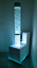 Image of a bubble tube featuring colorful lights and a flowing, water-like effect, set on a white podium. 