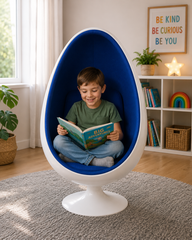 A young boy sits cross-legged inside a white egg-shaped chair with a blue cushion, reading a book in a bright, modern room with a bookshelf and plants in the background.