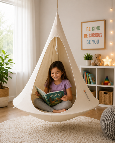 A child sits inside a cream fabric teardrop hanging chair in a bright, modern room, reading a book. The chair is suspended from the ceiling, and the background features a white bookshelf, a framed 'BE KIND BE CURIOUS BE YOU' sign, and a cozy atmosphere.