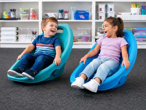 Two children sitting on bright blue and teal rocking chairs in a classroom setting with shelves of supplies in the background.