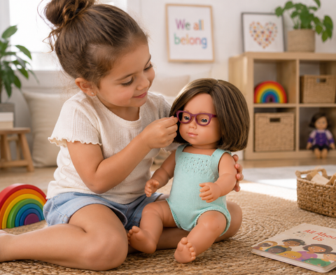 A young girl sits on a rug in a bright children's room, adjusting purple eyeglasses on a doll wearing a light blue romper. The room has a wooden shelf with toys, framed wall art, and a rainbow toy nearby.