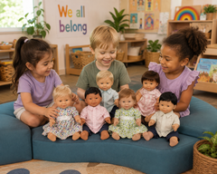 Three children sitting on a blue cushioned bench in a children's playroom, holding and playing with seven baby doll figures of different skin tones and outfits.