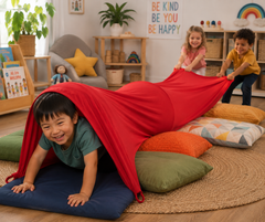 Three children playing with a red fabric tunnel made from a large sheet, supported by colorful floor cushions in a bright, playful children's room.
