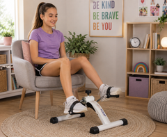 A woman sits on a gray armchair using a white and black pedal exercise bike in a bright, modern home office setting. The bike has the brand name 'SoleFit' visible on its frame.