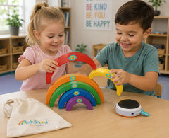 Two children playing with a Takiplay Rainbow wooden toy set on a light wood table in a classroom setting. The set includes five colorful wooden rainbow arches, a small white and blue magnetic tracking device, and a cream drawstring bag with the Takiplay logo.