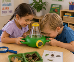 Two children, a girl in a purple shirt and a boy in a blue shirt, looking closely at a green and clear plastic bug observation kit on a wooden table in a classroom setting.
