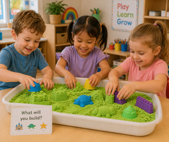 Three children are happily playing with green kinetic sand in a white tray, using colorful molded shapes and a small sign that reads 'What will you build?'. The scene is set in a bright, colorful children's classroom.