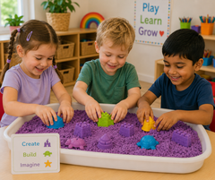 Three children play with a purple sand tray table in a bright classroom. The white plastic tray is filled with purple sand and colorful toy structures, with a small sign on the front that reads 'Create Build Imagine'.