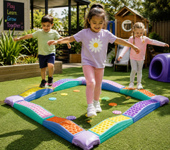 Three children playing on a colorful outdoor sensory balance mat on artificial grass in a playground setting.