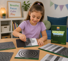 A young girl with a ponytail sits at a wooden table, actively playing with the 'Discover Sensory Cards' board game. The scene is set in a cozy, well-lit room with shelves, books, and a plant in the background.