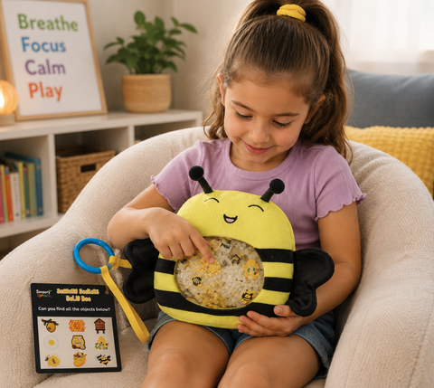 A young girl sits in a beige lounge chair in a cozy room, holding a bumblebee sensory toy. The toy has a yellow and black striped body with a clear belly showing gold glitter and confetti, and a blue ring puller.