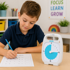 A young boy in a navy blue polo shirt sits at a wooden desk, writing on a worksheet with a blue pencil. Next to him is a white and blue digital timer.