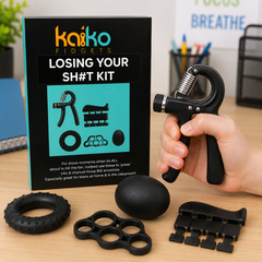 A hand holds a black spring-loaded hand grip trainer next to its retail box on a desk. The box reads 'KaKo Fidgets Losing Your SH*T Kit' and shows the product set, including the grip, ring, ball, and other black fidget accessories.