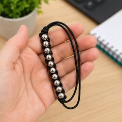 A black cord bracelet with silver metallic beads, held in a person's hand over a light wood desk background with a blurred plant and notebook.