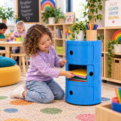 Child retrieving coloured paper from a blue Componibili storage unit in a vibrant preschool classroom.