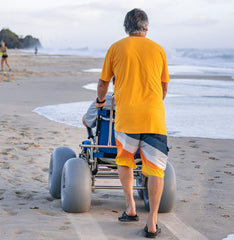 This is an image of the Sandcruiser All Terrain Dune Buster Wheelchair. It shows a person being pushed along the beach by another person. It is Blue and has two large WheelEEZ wheels on the front and two large WheelEEZ wheels on the back.