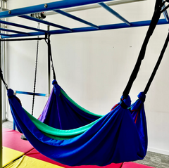 A colorful layered Lycra therapy hammock hangs from a horizontal ladder above, showcasing a purple top layer with blue, green, and red layers beneath.