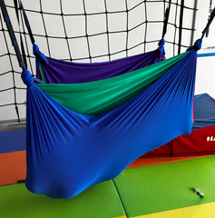 Colorful layered Lycra therapy hammock featuring a purple top layer with blue, green, and red layers beneath with a person inside, hanging from a net structure.