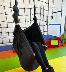 A black layered therapy swing hammock hangs from the ceiling, with a person sitting inside and their legs hanging outside.