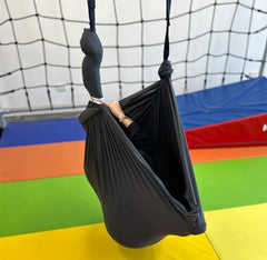 A black layered therapy swing hammock hangs from the ceiling, with a person sitting inside 