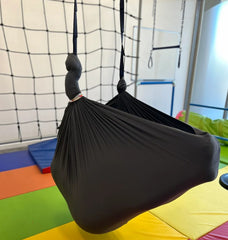 A black layered therapy swing hammock hangs from the ceiling, with a person sitting inside with a net background.