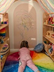 Young girl lies in her play nook with a blush coloured Magnetic Chalk Wall Archway that encourages creative expression and mark-making.