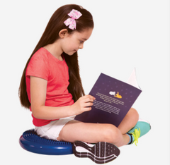 A young girl sits cross-legged on the floor, perched on a blue tactile cushion, reading a book.
