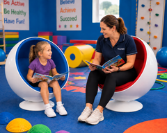 A child sits in a blue ball chair reading a book, while a teacher relaxes in a red ball chair.