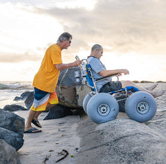 This is an image of the Sandcruiser All Terrain Dune Buster Wheelchair. It shows a person being pushed over sandbags and rocks at the beach by another person. It is Blue and has two large WheelEEZ wheels on the front and two large WheelEEZ wheels on the back.