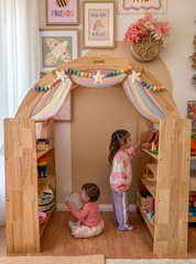 This is an image of the Wooden Interactive Play Stand and Retreat Den with colorful fabric and toys on the shelves. Two children are playing inside.