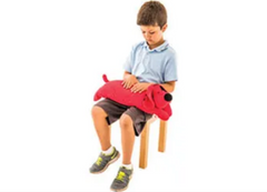 Image of a child sitting on a chair with lap pad shaped like a dog. It is a red dog with a black nose.