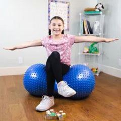 This is an image of a girl sitting on a blue Therapy Inflatable Sensory Ball with Tactile Nubs that  is a peanut shape. She has her arms outstretched. It has tactile nubs  on the outside.